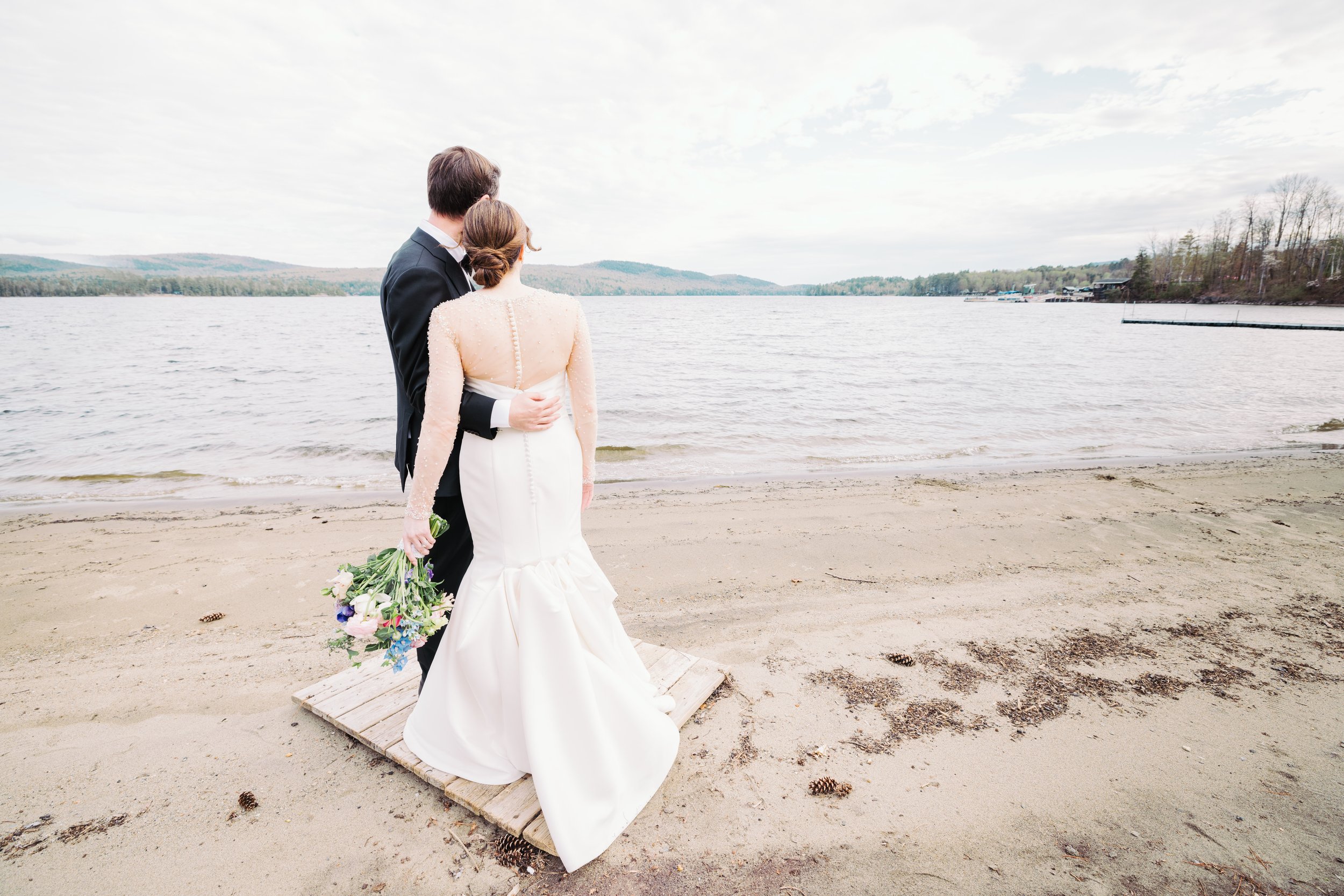 A bride and groom stand on a small wooden platform on a beach, facing a lake with hills in the background, during daytime.