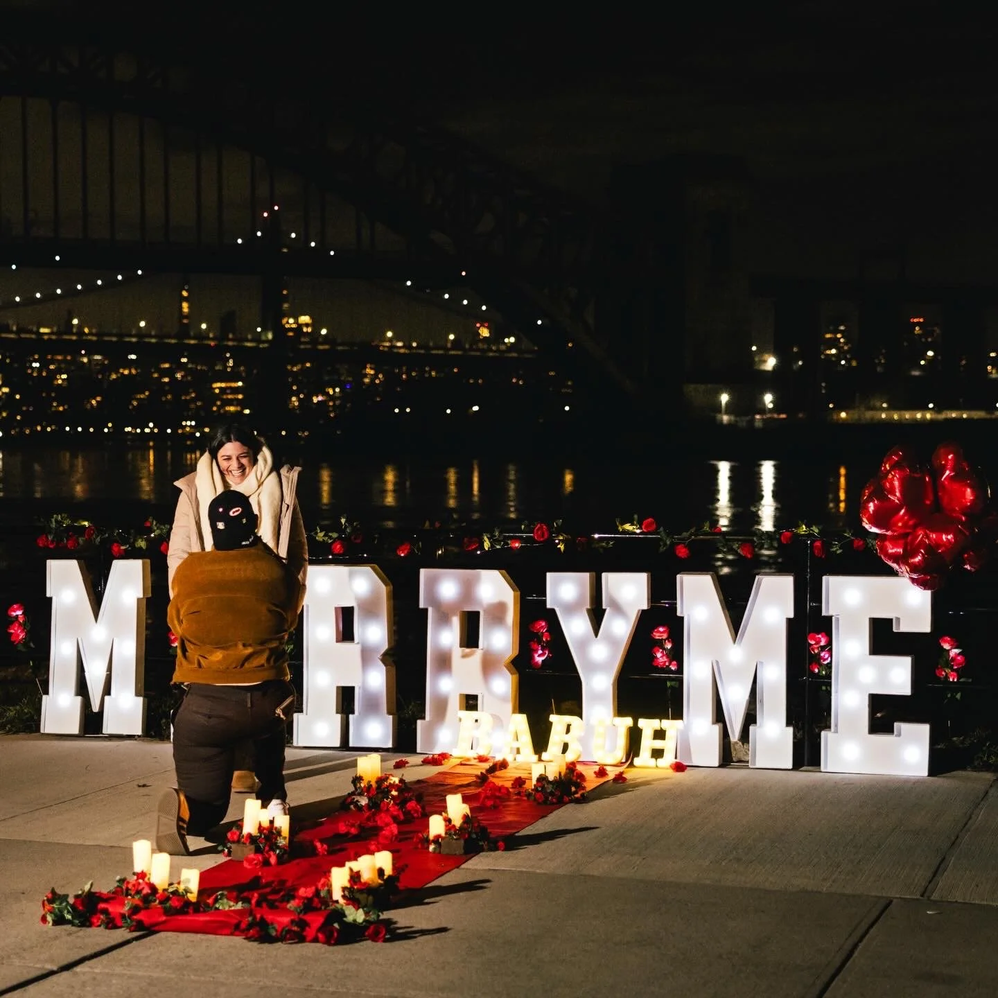 A couple celebrating a marriage proposal on a decorated walkway with illuminated letters spelling 'MARRY ME' and 'BABY' at night near a bridge and city skyline.