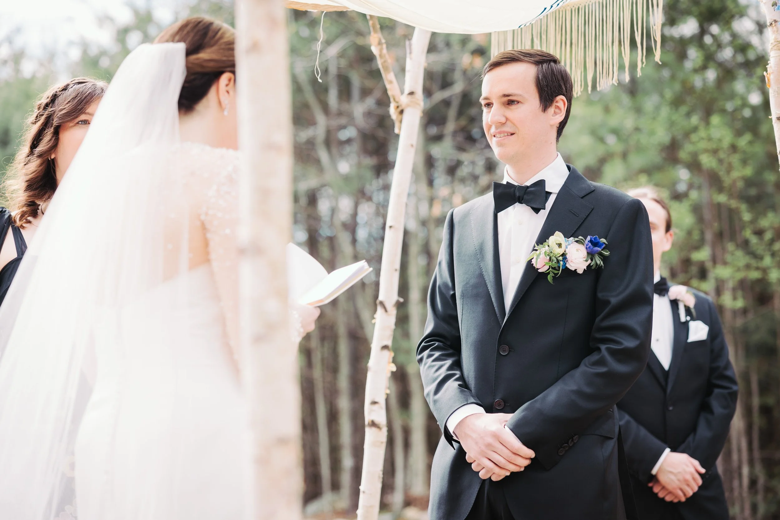 A man in a black tuxedo and a woman in a wedding dress with a veil exchanging vows at an outdoor wedding ceremony.