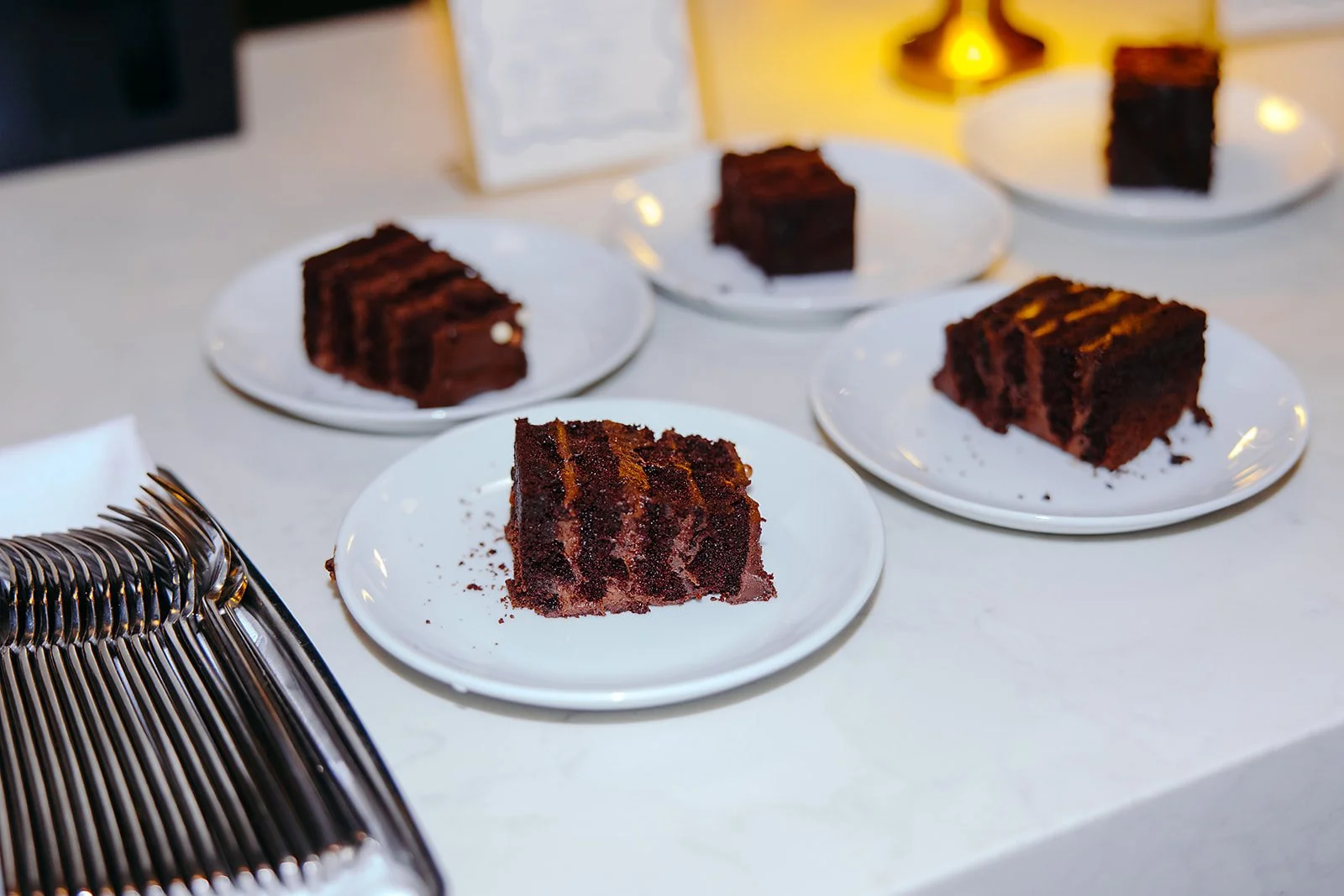 Slices of chocolate cake on white plates on a white table, with a cake display in the foreground and warm lighting in the background.
