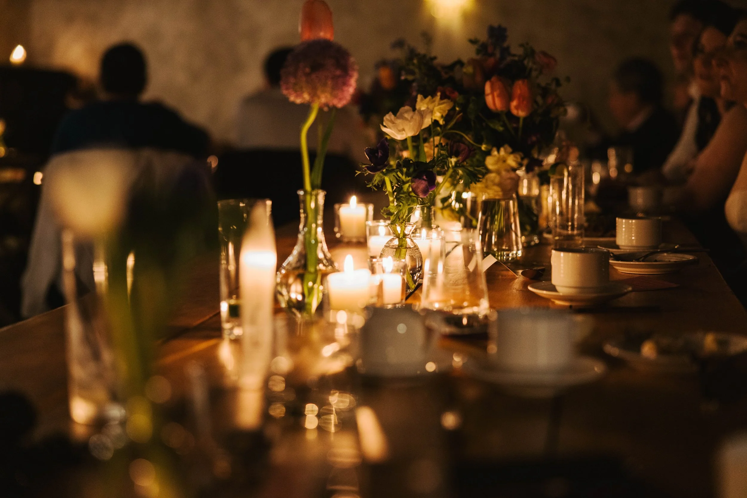 A dimly lit dinner table with candles and floral centerpieces, with people seated around it.