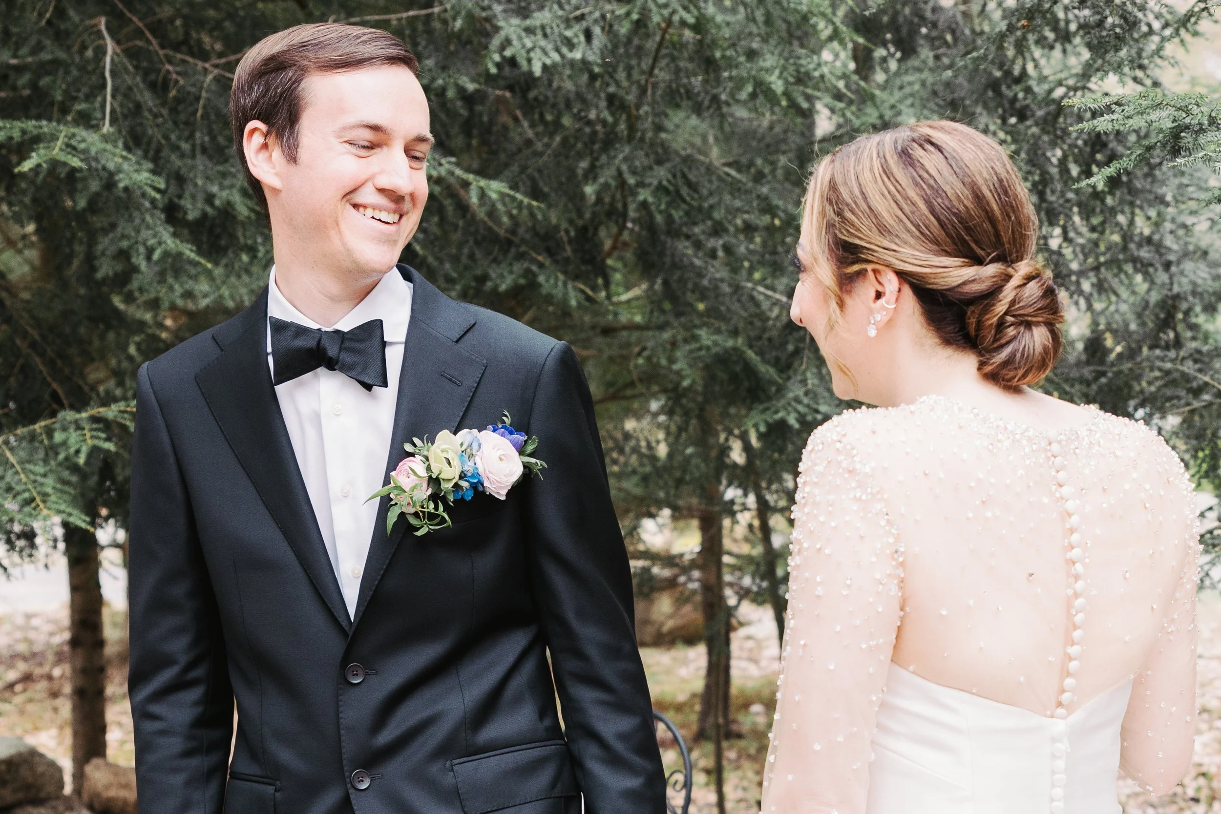 A groom in a black tuxedo with a bow tie and boutonniere, smiling at a bride with her back to the camera, wearing a white wedding dress with pearl embellishments, outdoors near green trees.