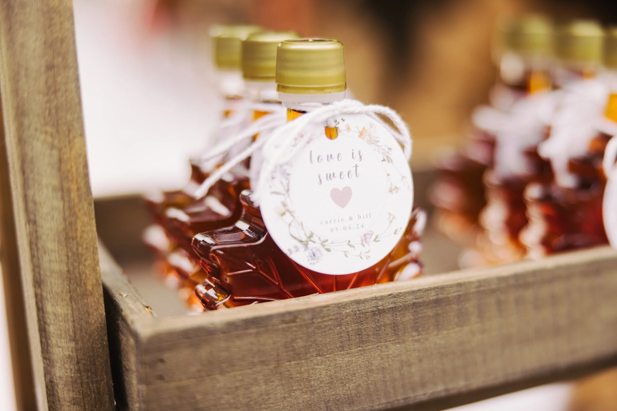 Small glass bottles filled with amber-colored honey, decorated with white tags that say 'love is sweet' and personalized with names and date, tied with white string, displayed in a wooden box.