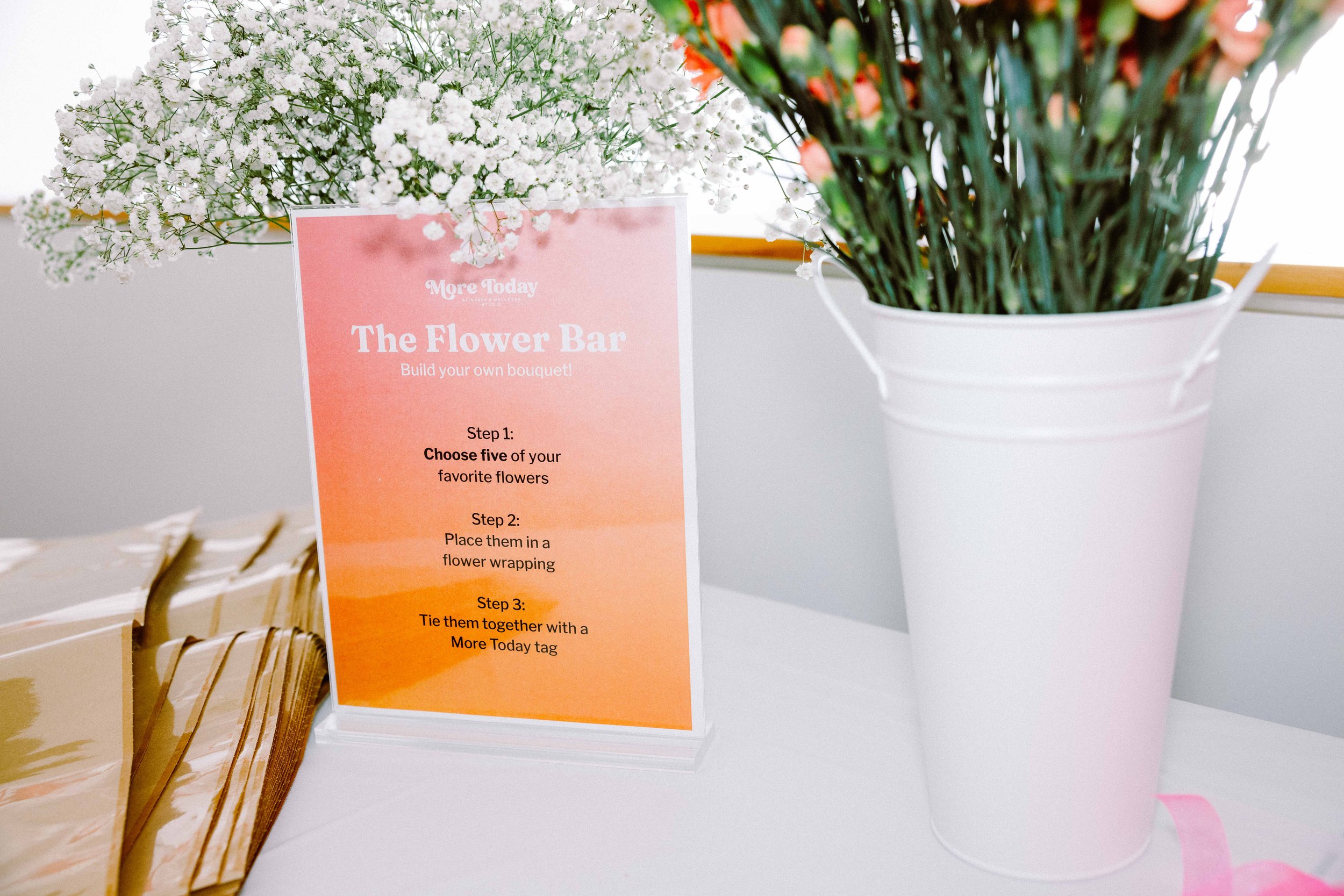 A sign titled 'The Flower Bar' details steps for creating a flower bouquet, with white flowers and pink flowers in vases beside it.
