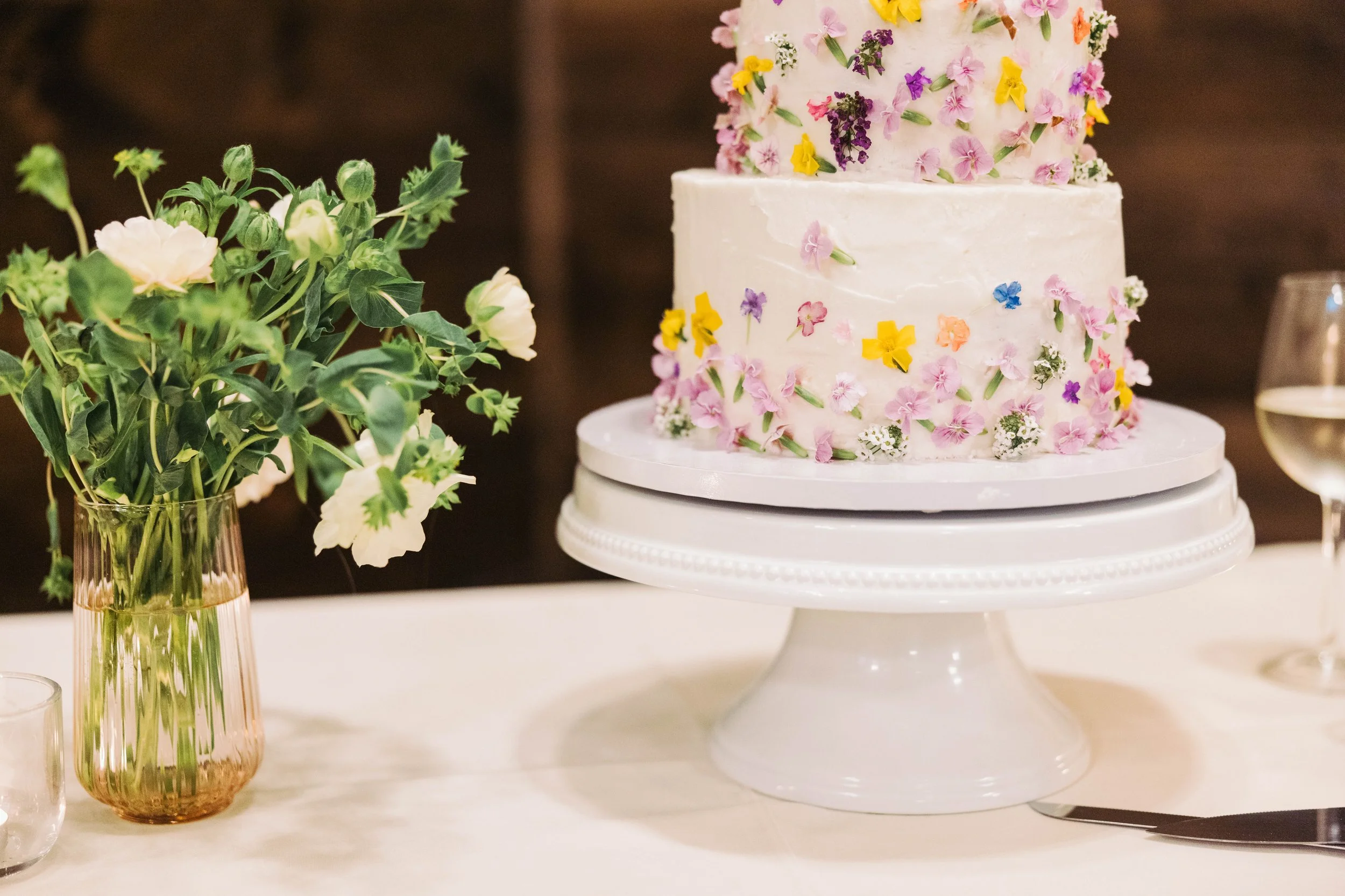 A three-tiered wedding cake decorated with small, colorful edible flowers, placed on a white cake stand. To the left, a tall glass vase holds a bouquet of white and green flowers and foliage. To the right, a glass of white wine is partially visible.