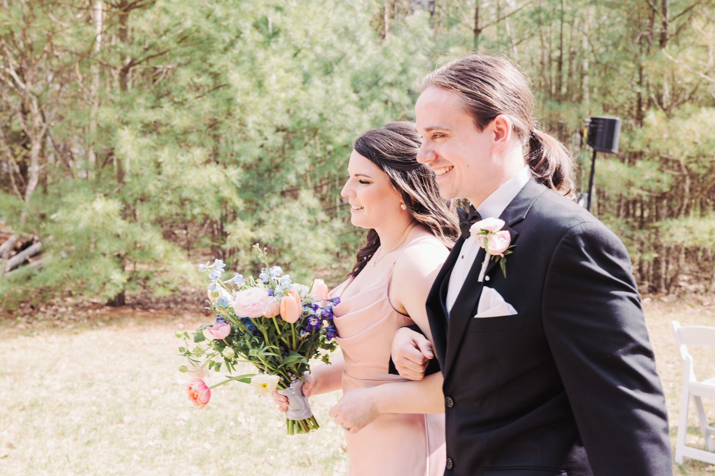 A bride and groom walking outdoors on their wedding day, smiling and holding hands. The bride is holding a bouquet of pink, purple, and white flowers, and the groom is wearing a black tuxedo with a pink boutonniere. The background features green tree