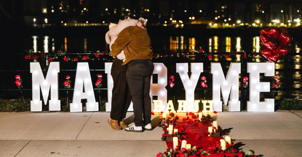 A couple hugging by a waterfront decorated with large illuminated letters spelling 'MARRY ME,' surrounded by flowers and candles for a marriage proposal.