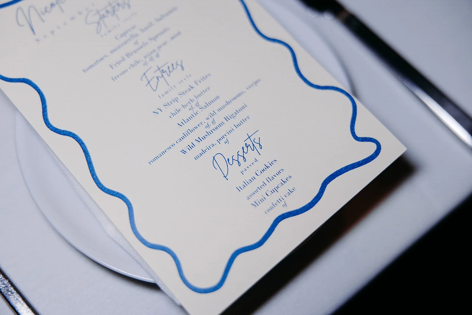 A printed menu or food list with a blue wavy border, listing dishes such as filled or stuffed items, mussels, cookies, cupcakes, and cake, served in a restaurant or event setting.