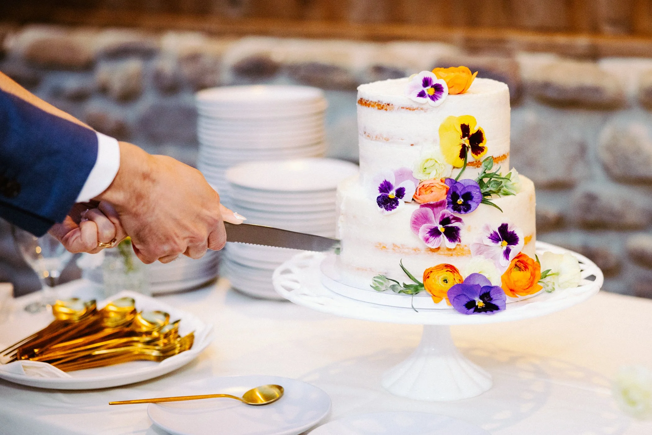 A person in a dark suit is cutting a two-tier white wedding cake decorated with colorful edible flowers on a white cake stand. Behind the cake are stacks of white plates. Gold utensils are on a plate to the left.