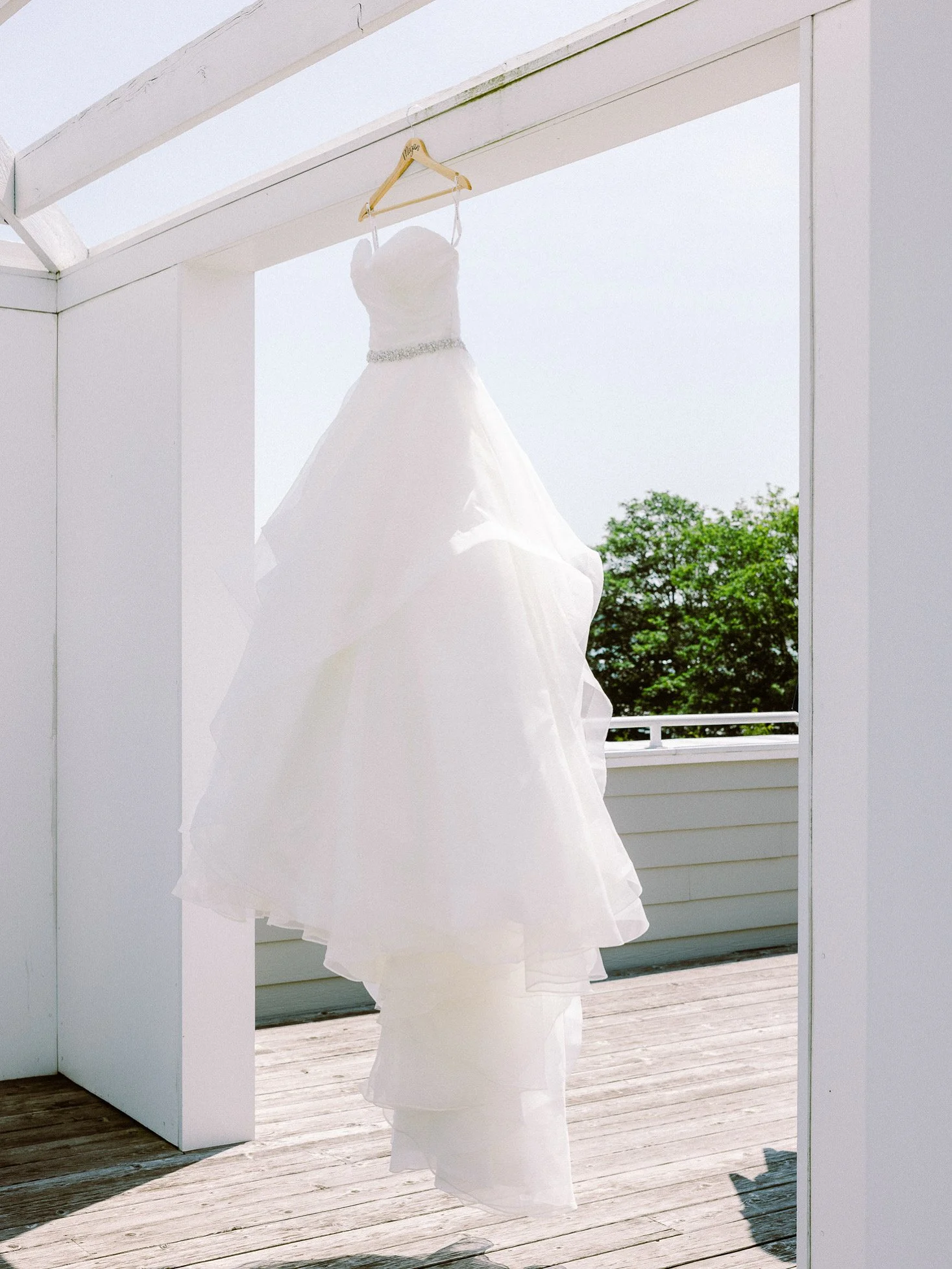 A white wedding dress hanging on a wooden hanger in an open frame structure, with trees and sky in the background.
