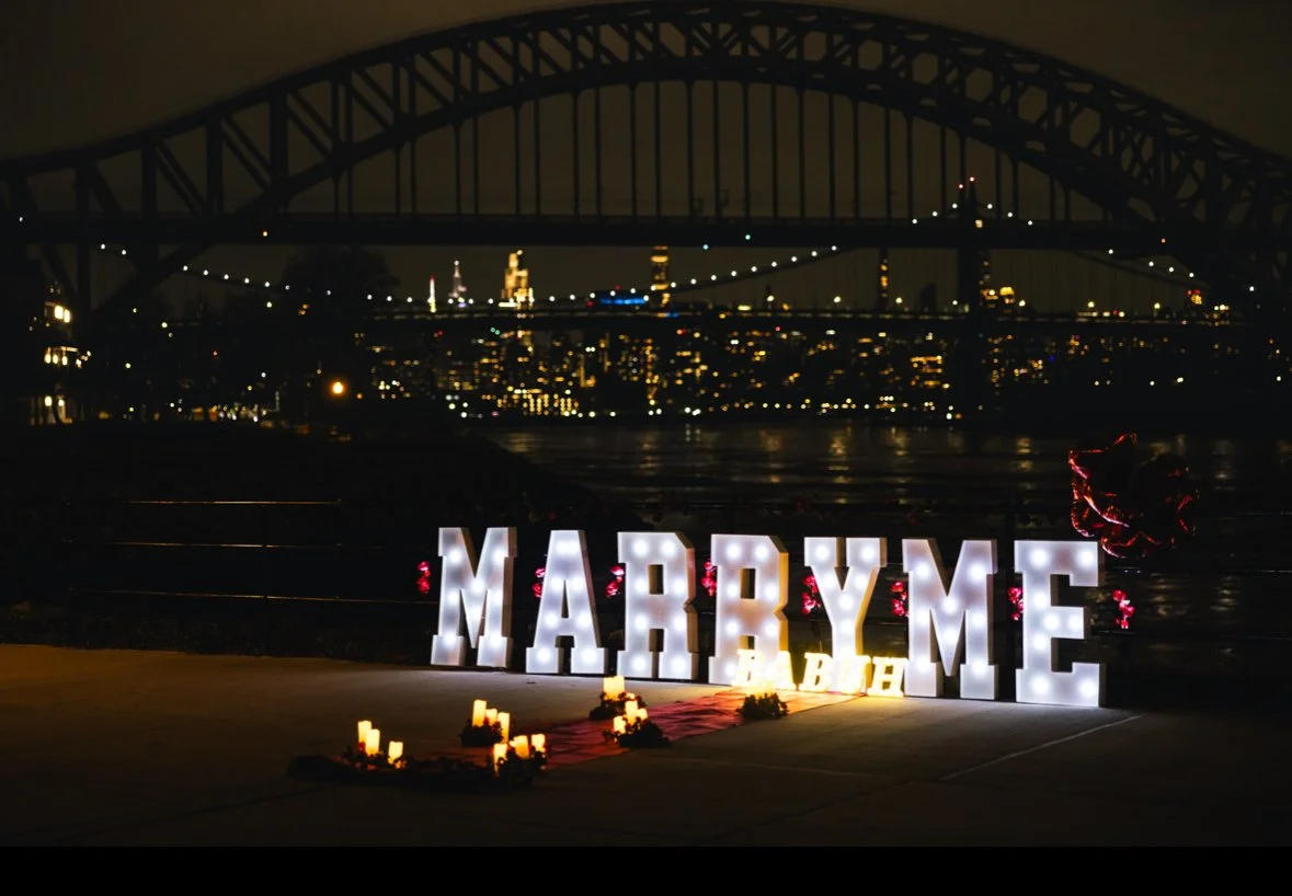 Large illuminated letters spelling "Marry Me" on a walkway near the water at night, with a bridge and city skyline in the background.