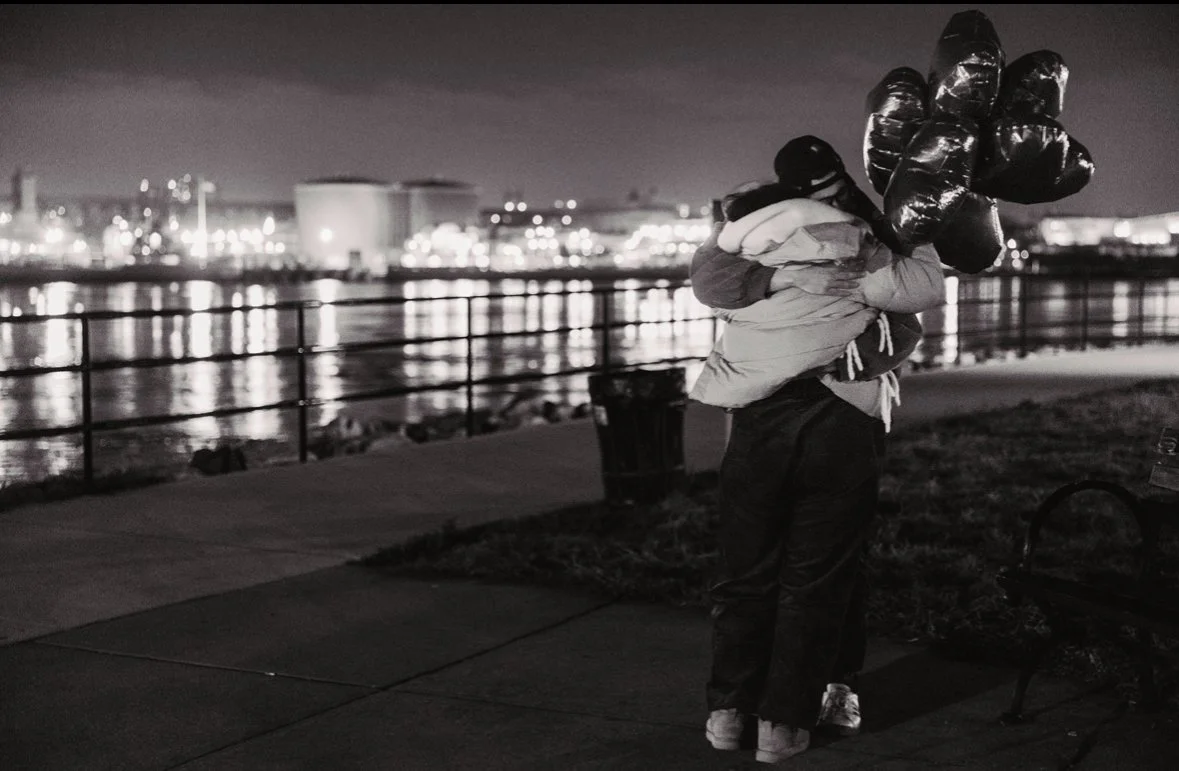 A person holding a bouquet of black balloons at night by a waterfront, with city lights reflecting on the water in the background.