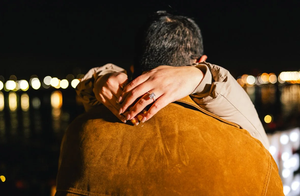 A person with short hair embracing another person at night, with city lights in the background.