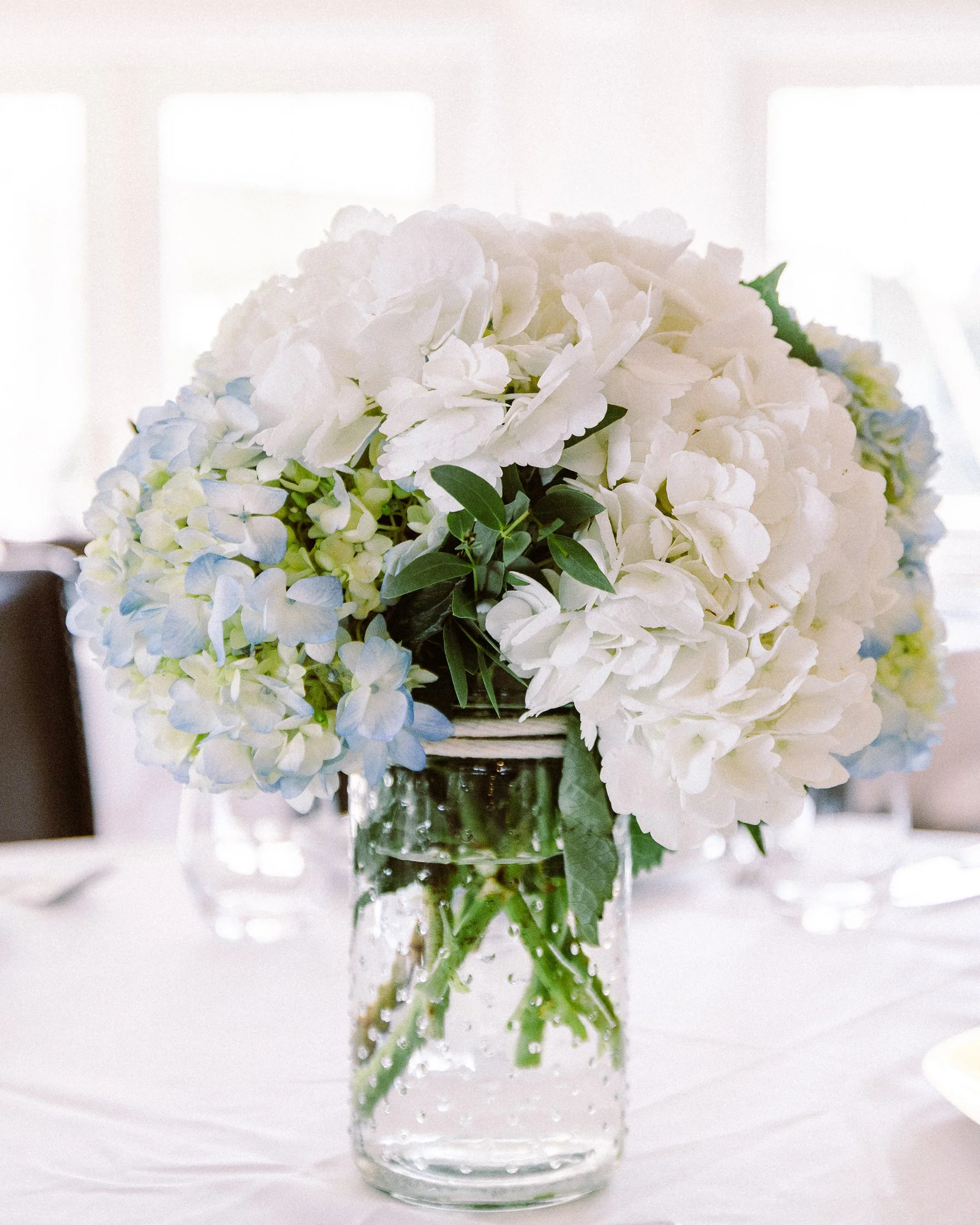 A bouquet of white and light blue hydrangeas in a clear glass vase on a white tablecloth.