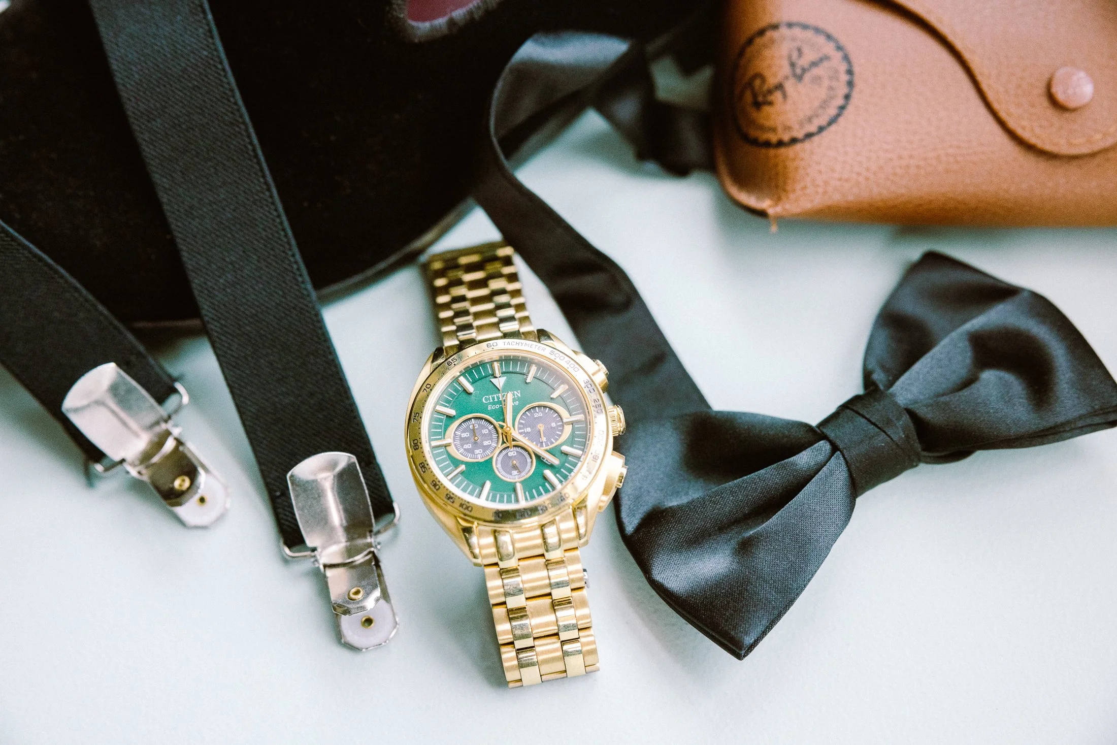 A gold Citizen wristwatch with a green face, three subdials, and a metal band, surrounded by suspenders, a black bow tie, and a brown leather case.