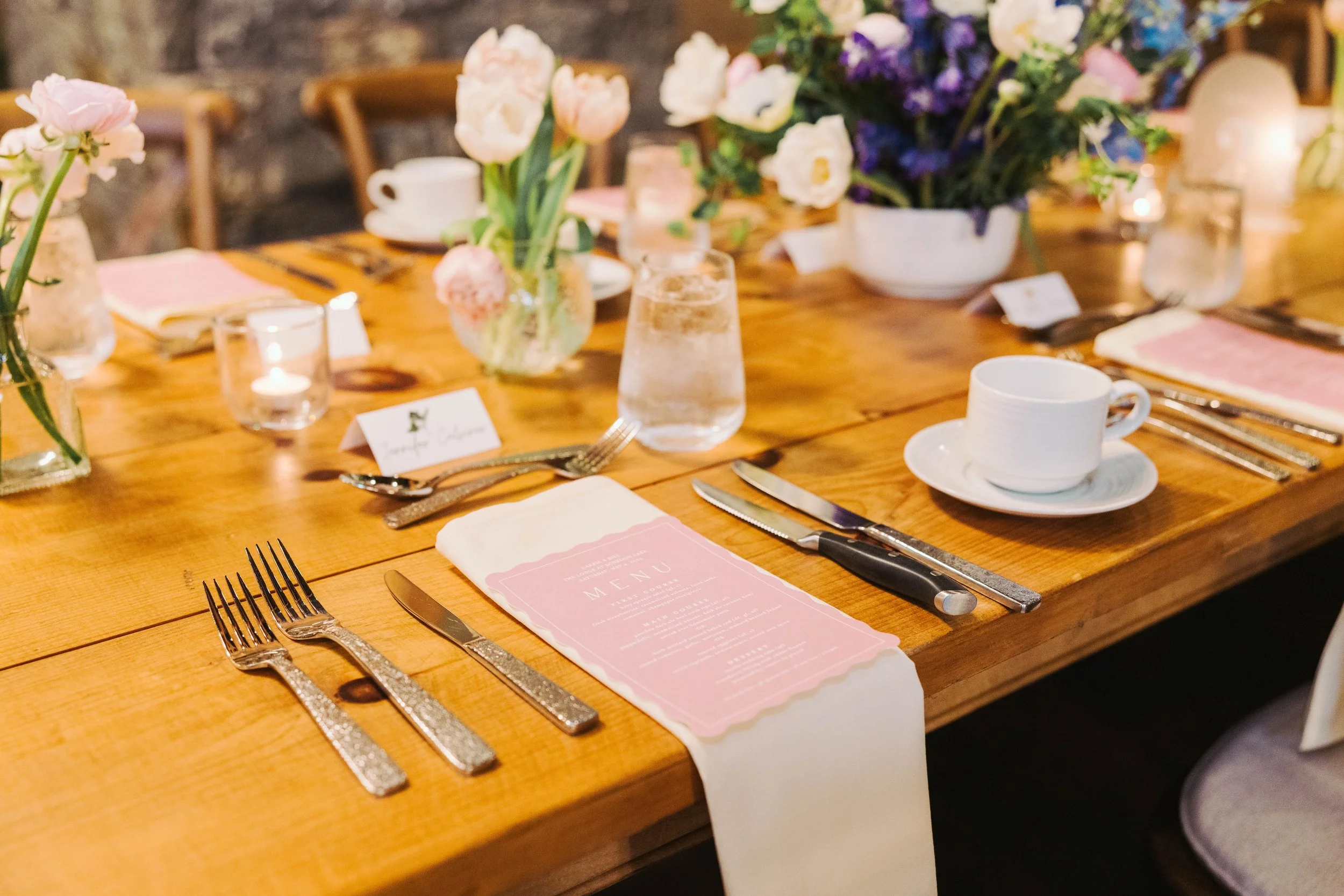 A beautifully set dining table with pink floral arrangements, a menu, tea cup and saucer, glasses of water, cutlery, and candles, decorated with flowers and elegant tableware.