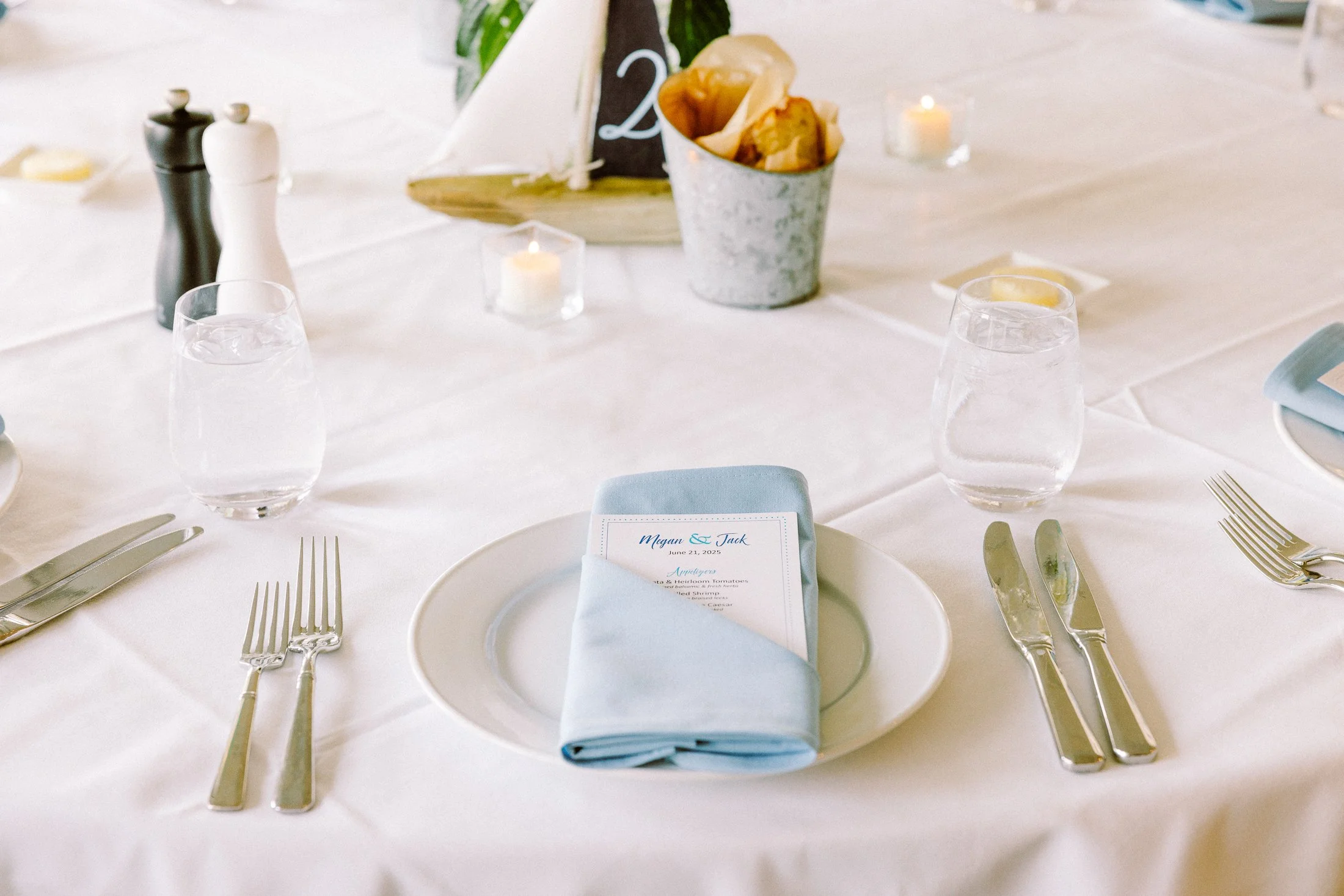 A formal dining table set for a wedding reception with a white tablecloth, divided into multiple place settings. Each setting has a white plate with a folded light blue napkin on top, with a printed menu inside. Silverware is neatly arranged with kni
