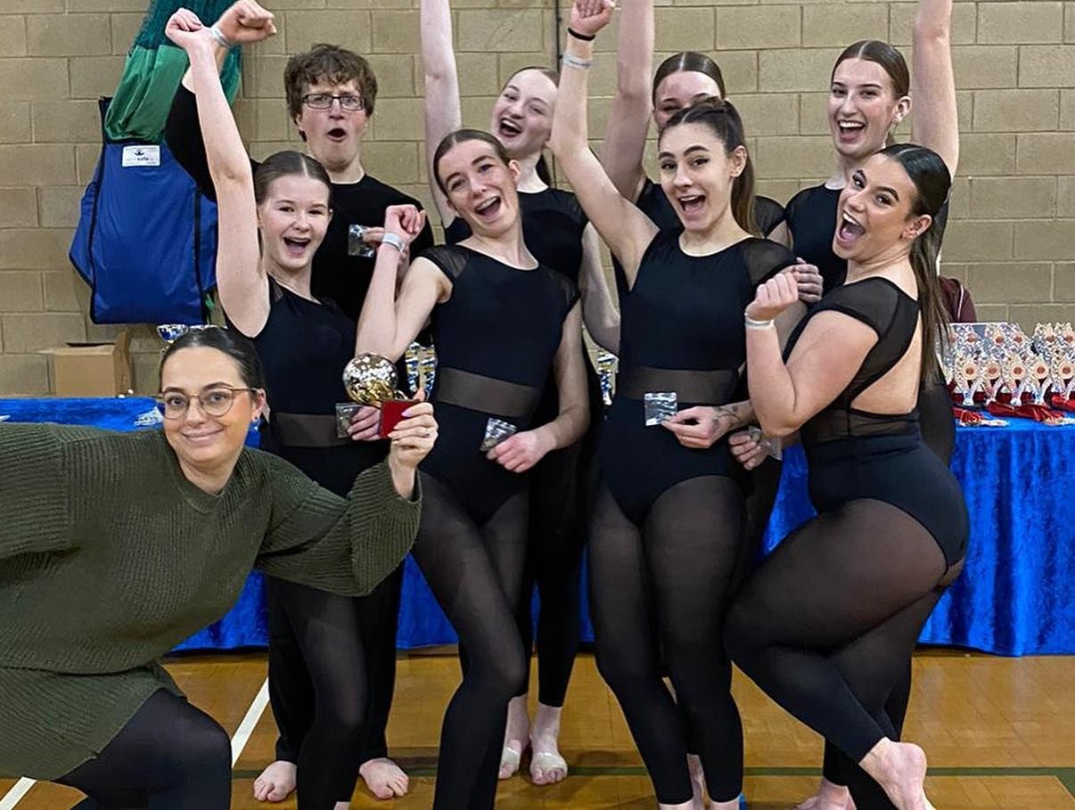 A group of eight young women and one young man celebrating indoors, with some women holding medals and showing victory poses, wearing black dance costumes and tights.