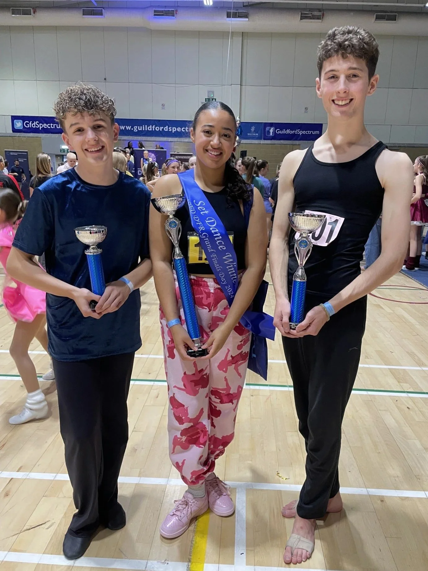Three young athletes standing on a gymnasium floor, each holding a trophy. The girl in the middle is wearing a blue sash and pink camouflage pants, smiling. The boys on either side are dressed in athletic wear, also smiling. There are people and banners in the background.