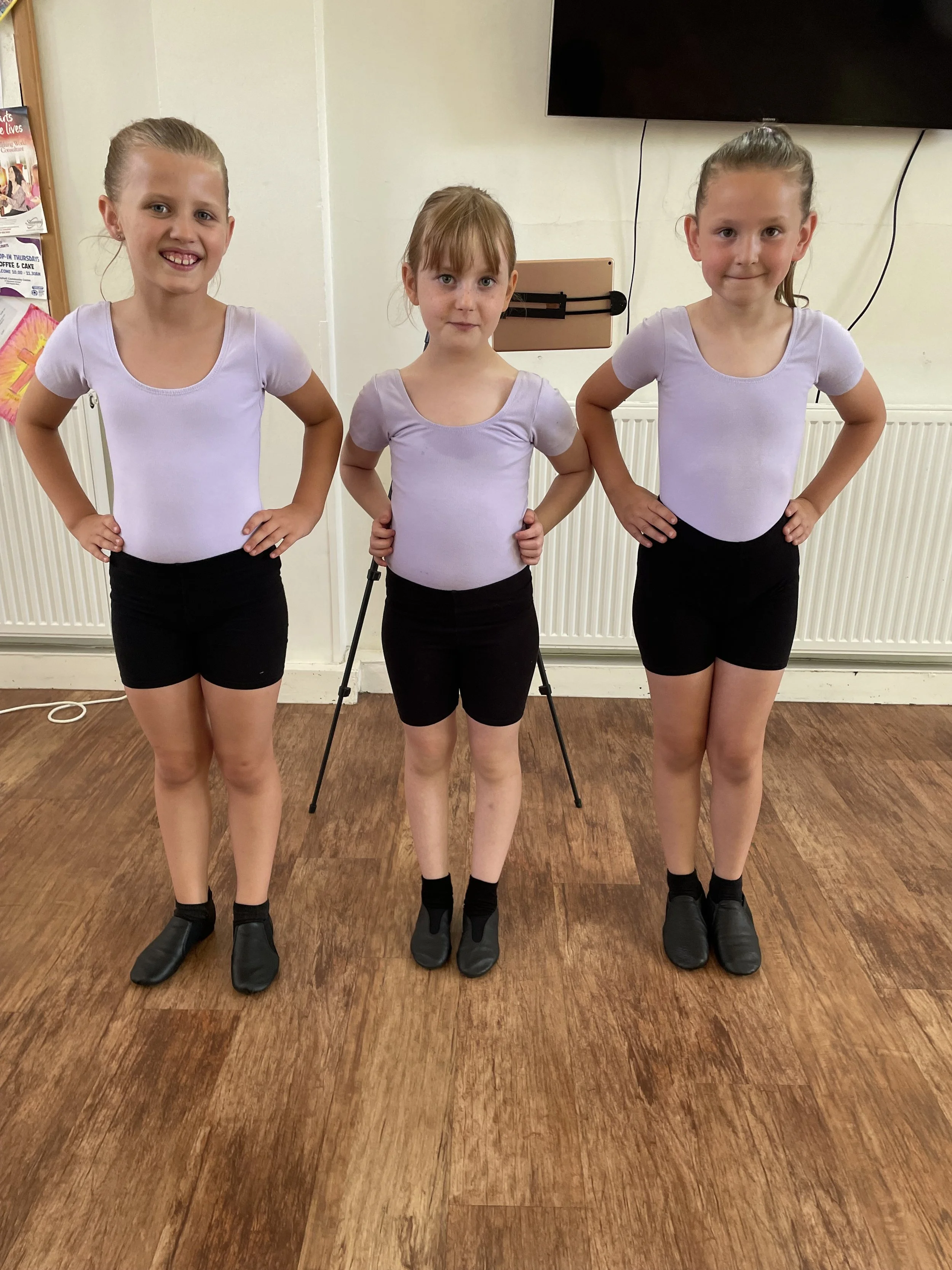 Three young girls in dance outfits standing in a line with hands on hips, on a wooden floor in a dance studio.