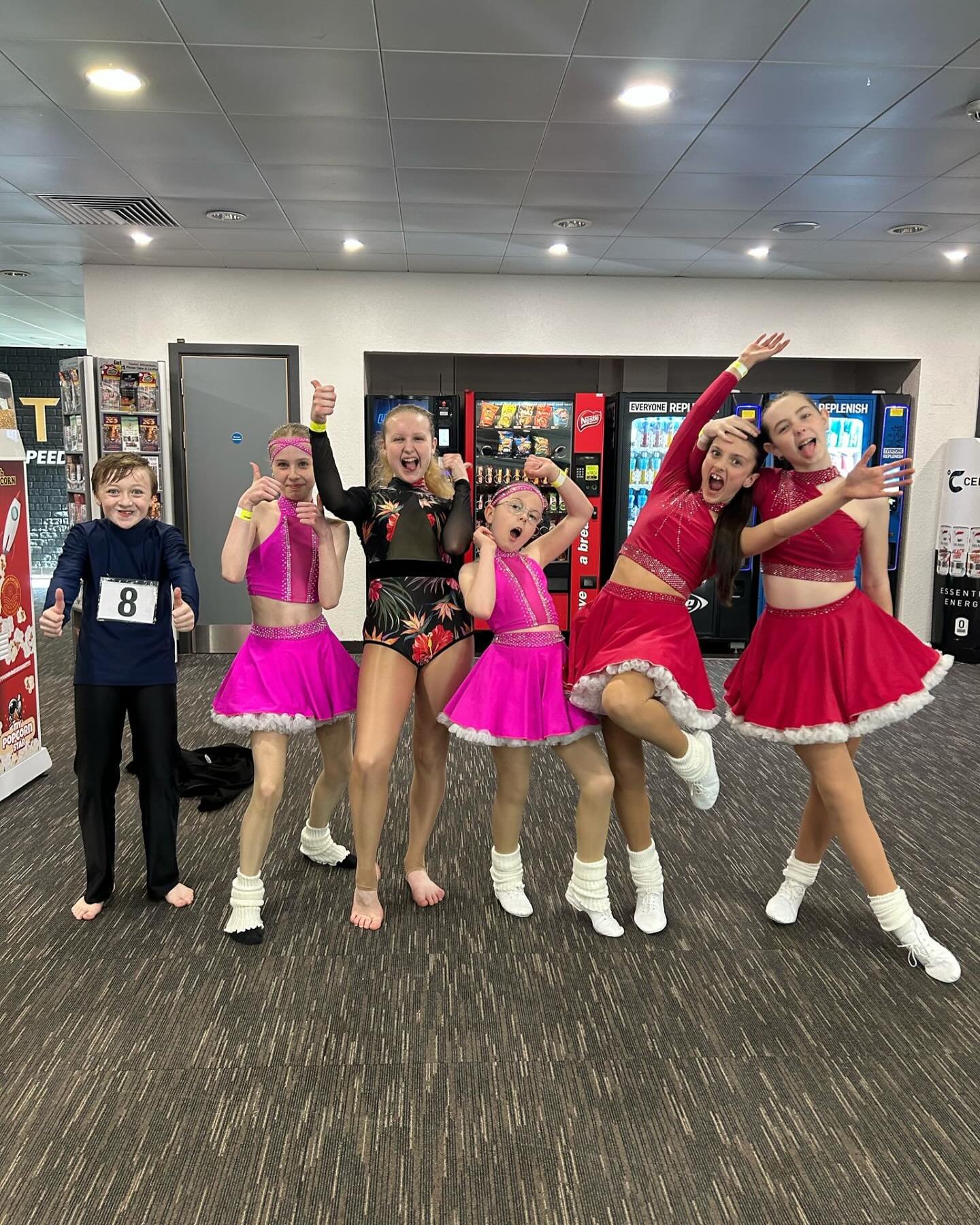Group of young dancers and a boy posing and cheering in an indoor setting, some in bright costumes with skirts and socks.