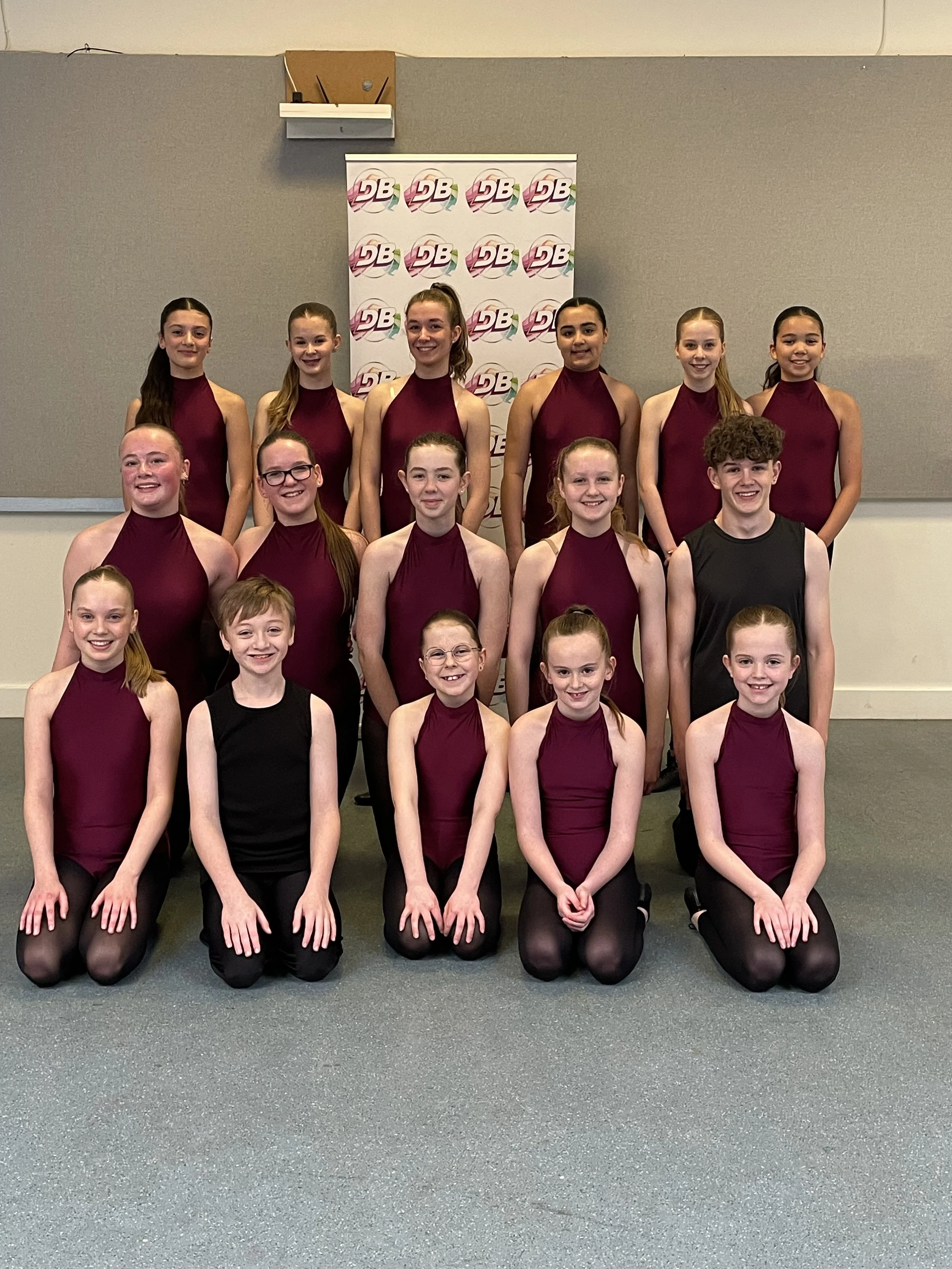 Group of young girls and one boy in matching dance costumes posing for a photo indoors in front of a backdrop with logo. All are smiling.