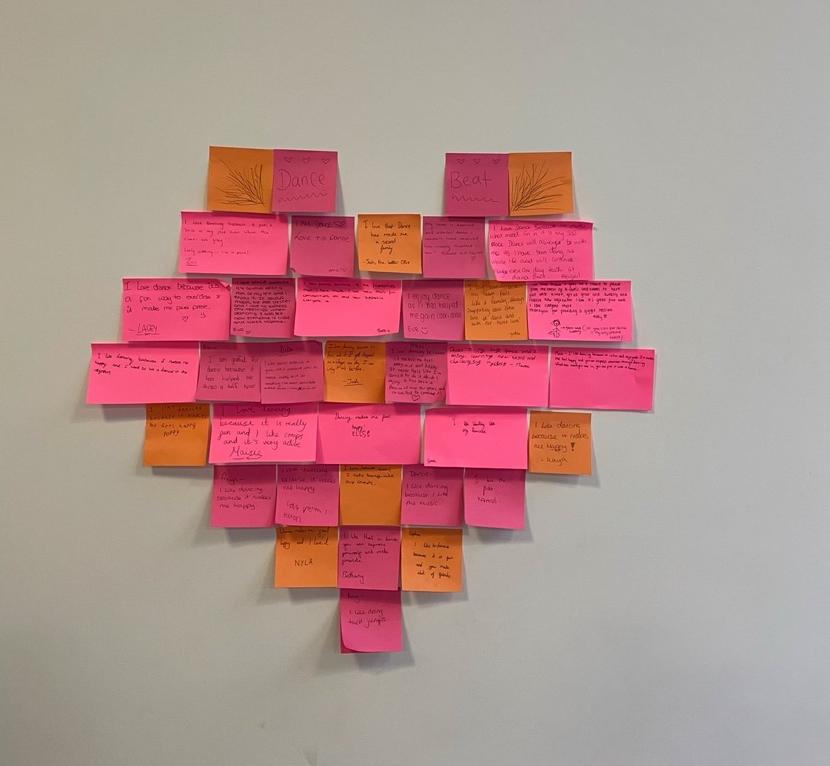 Heart-shaped collage of colorful sticky notes with handwritten notes and drawings on a plain white wall.
