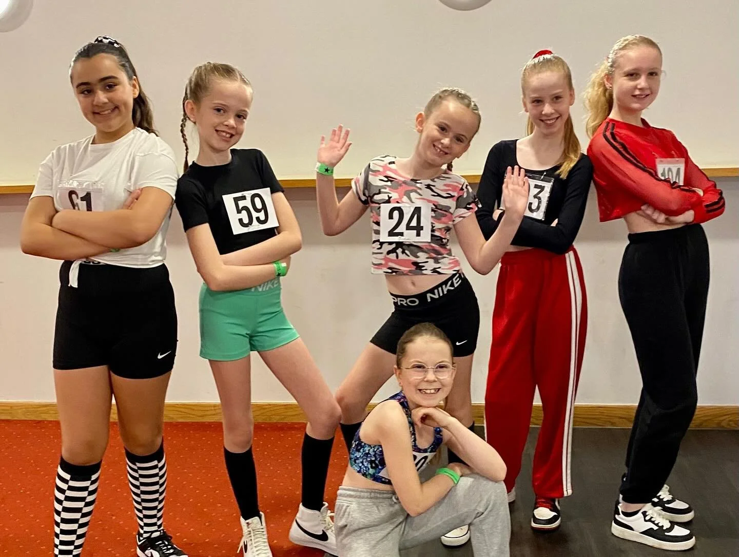 Group of six young girls, one sitting on the floor and five standing, dressed in athletic clothing, smiling and posing at an indoor event.