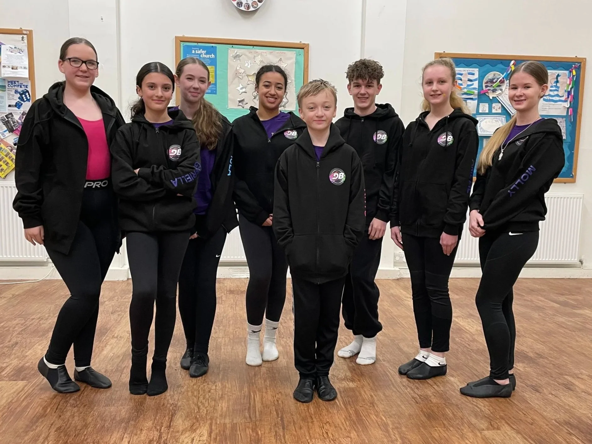 Group of nine young people standing indoors on a wooden floor, wearing black hoodies and black pants, with some in athletic clothing, in front of a board with posters and decorations.