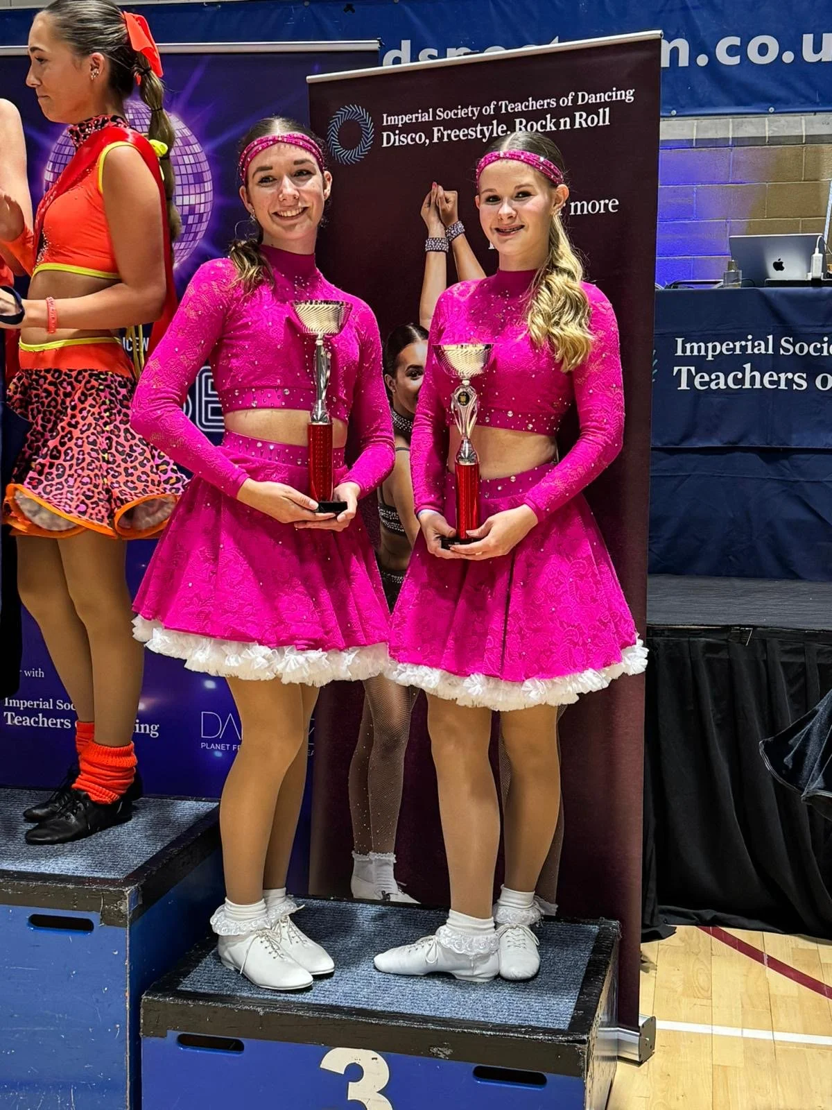 Two young women in matching bright pink dresses, holding awards, standing on a winners' podium at a dance competition.