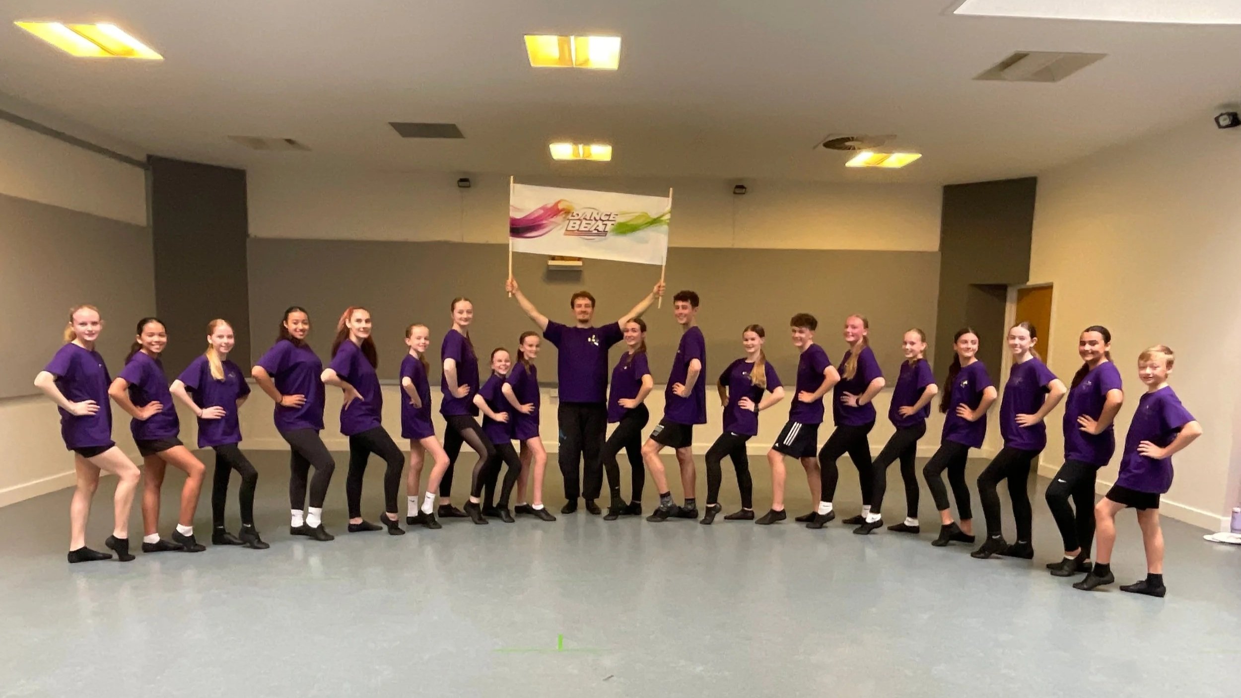 A group of young girls in purple shirts and black shorts or leggings standing in a line in an indoor room, with a man in the center holding a flag with the logo 'Dance Beat' above his head. The girls are posing with hands on hips, smiling.