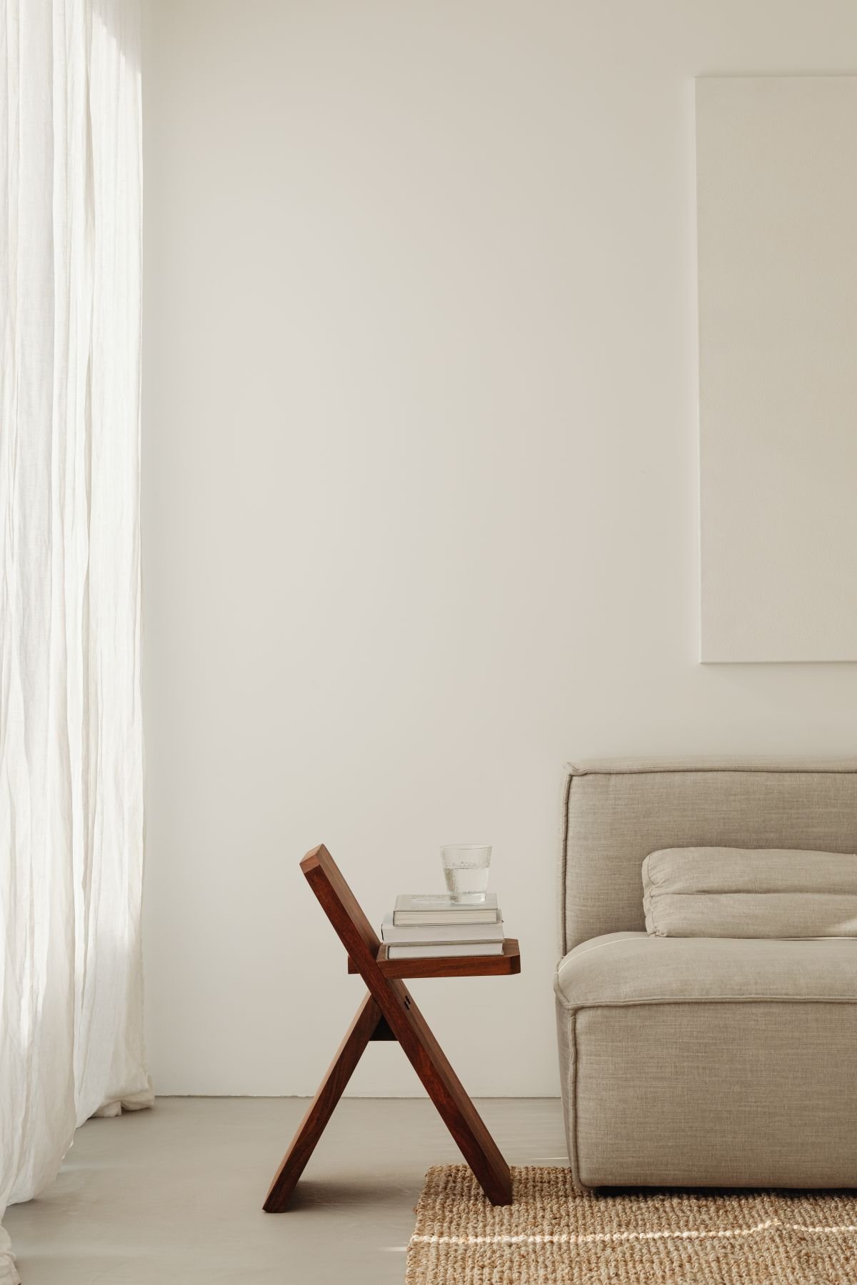 Custom Squarespace Template, E-commerce Squarespace Template, Avelã Creative. Minimalist living room with a beige sofa, wooden side table with books and a glass of water, sheer curtain, and woven rug on the floor.
