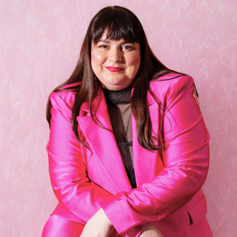 A woman with long dark hair, wearing a shiny pink blazer over a black top, sitting against a pink patterned background, smiling at the camera.