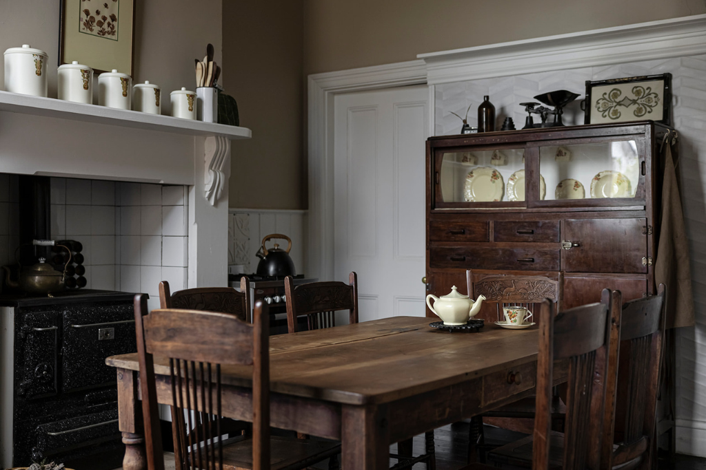 A vintage kitchen with a wooden dining table, six matching chairs, a wooden cabinet with glass doors displaying dishes, a black wood-burning stove with a tea kettle, and a white teapot with a cup on the table.