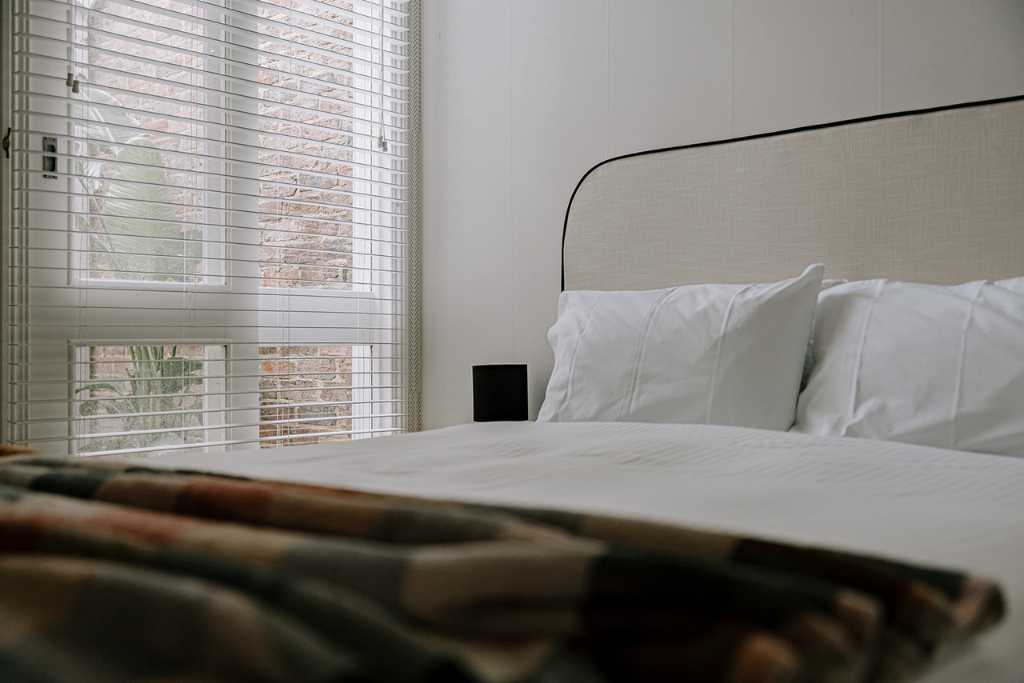 A neatly made bed with white pillows and sheets, a beige headboard, a black nightstand, and a window with white blinds showing a brick wall and outside greenery.