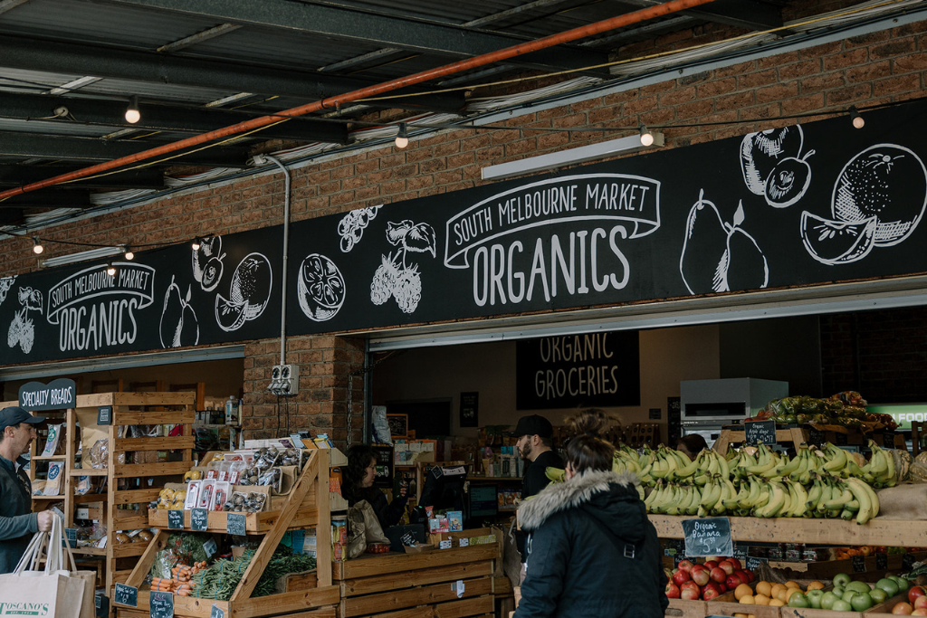 Indoor view of South Melbourne Market's organic produce section with fresh fruits and vegetables, and customers shopping.