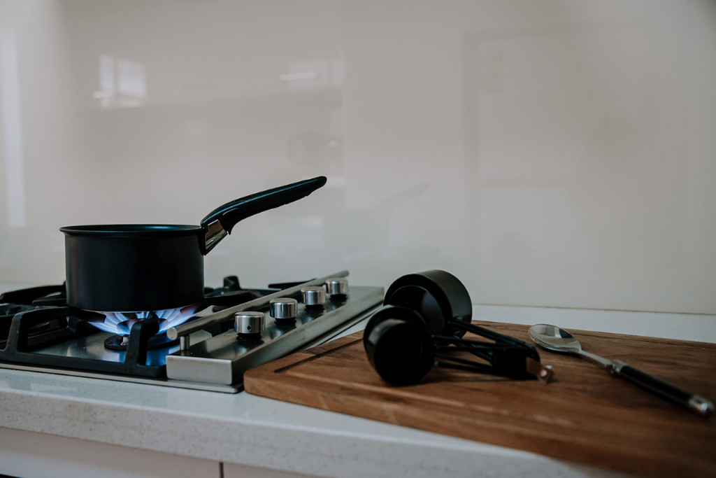 A black saucepan on a gas stove with a blue flame, a wooden cutting board, a set of black measuring cups, and a silver spoon on the countertop.