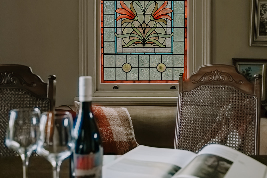 Interior of a dining room with a stained glass window, wooden dining chairs, a bottle of wine, two wine glasses, a book, and framed pictures on the wall.