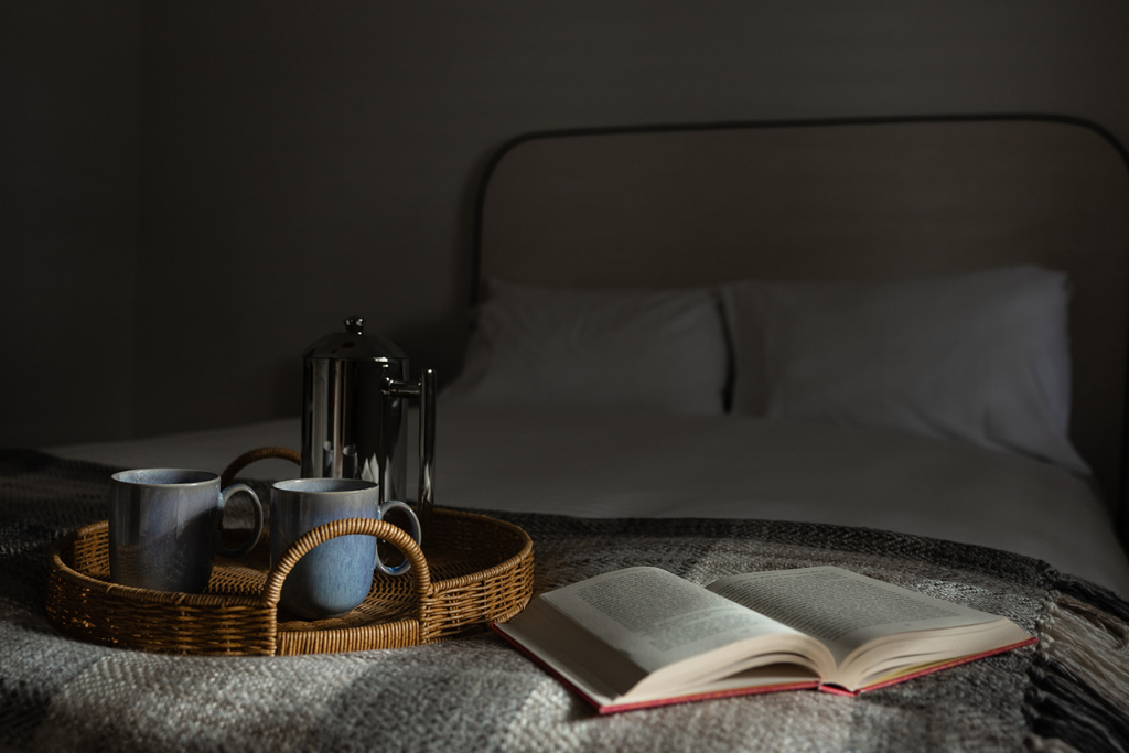 An open book, two coffee mugs, and a French press on a tray on a bed with white pillows in a dimly lit room.