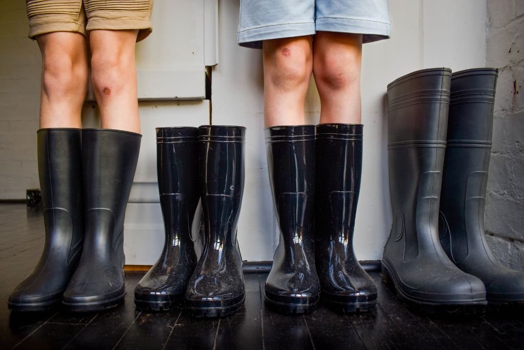 Two children standing in gum boots on a dark wooden floor in front of a white wall with a door.