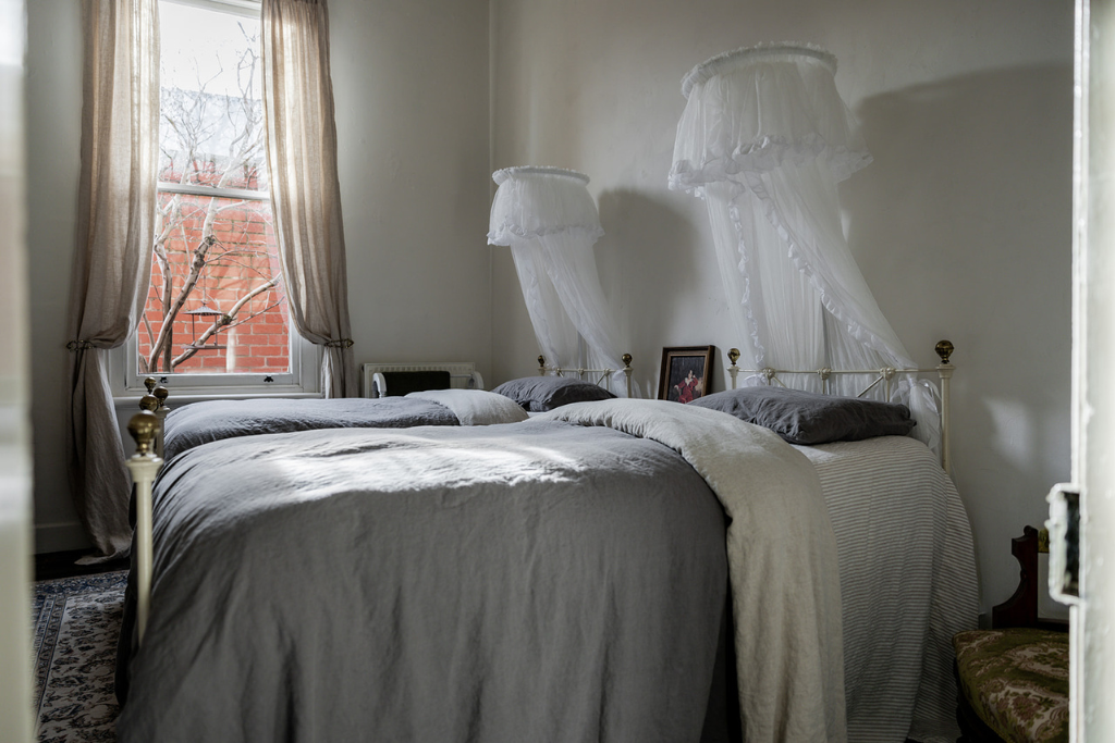 A bedroom with two beds covered in gray and white bedding, two canopy-like mosquito nets hanging above, a window with beige curtains, and a framed portrait on the bed's headboard.