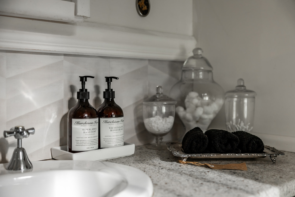 Bathroom countertop with two brown bottles of hand soap and hand cream, jars with cotton balls and cotton swabs, and a tray with black rolled towels.