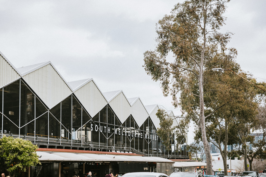 Exterior view of South Melbourne Market with large trees in the foreground and market signage visible on a modern building with metal and glass facade.