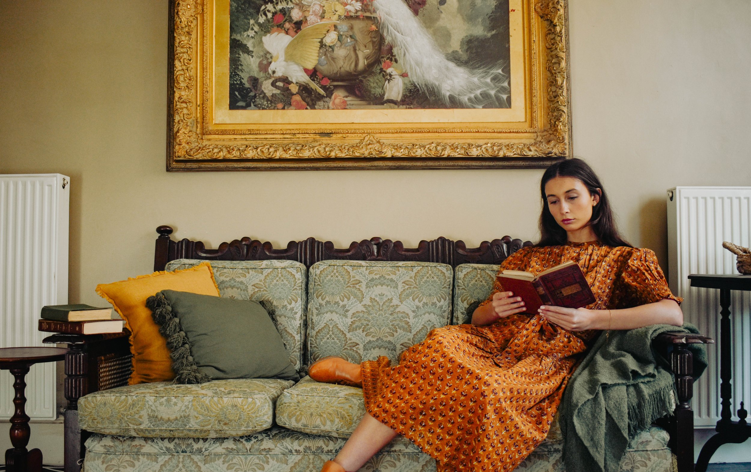 A young woman with dark hair sitting on a vintage-style sofa, reading a book. She is wearing a brown patterned dress. The sofa has a yellow and green pillow, and there are books on a small table beside her. A large framed painting of a bird and flora