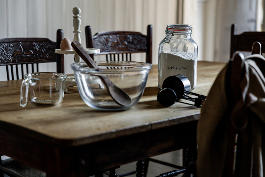 A wooden dining table set with a glass jar labeled 'Self-Rising Flour', a glass mixing bowl with a spatula, a small glass measuring cup, a pair of black kitchen scales, and a beige apron hanging on a chair, with carved wooden chairs around the table.