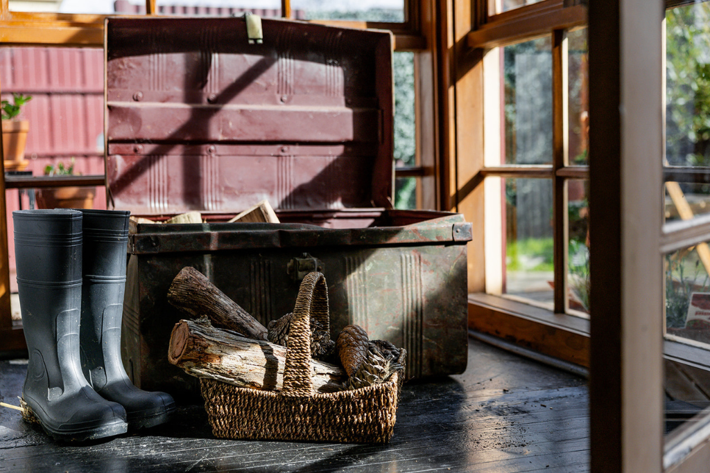 A pair of black rubber boots, a woven basket with firewood, and an open red metal chest in a sunlit indoor space with wooden framed glass doors and a garden view.