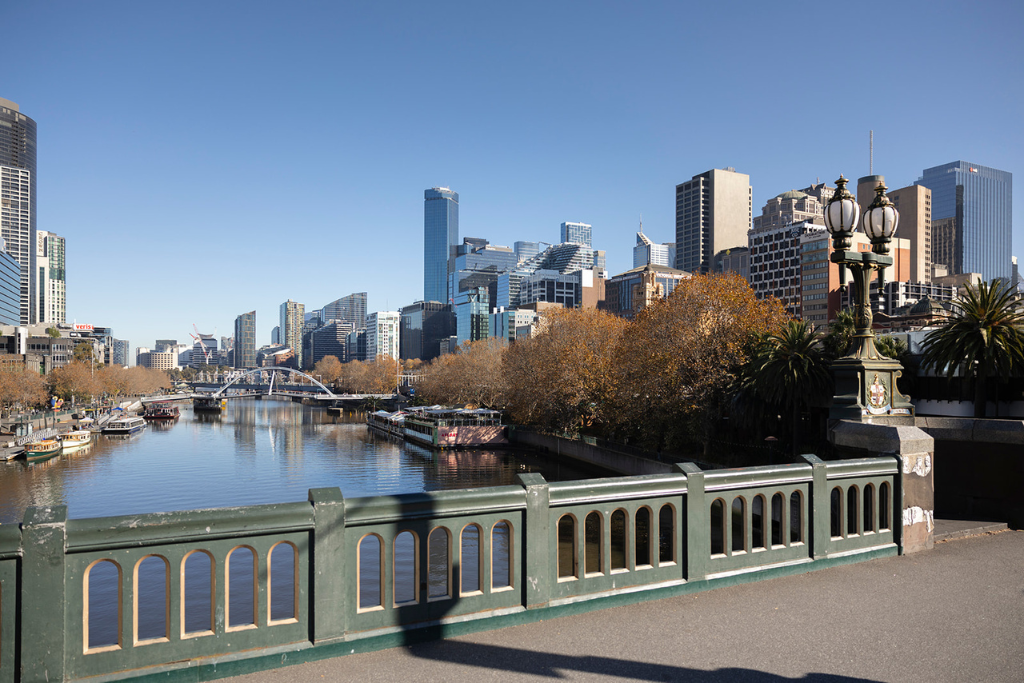 City skyline with tall skyscrapers across the Yarra River, with trees along the riverbank and a decorative bridge in the foreground.