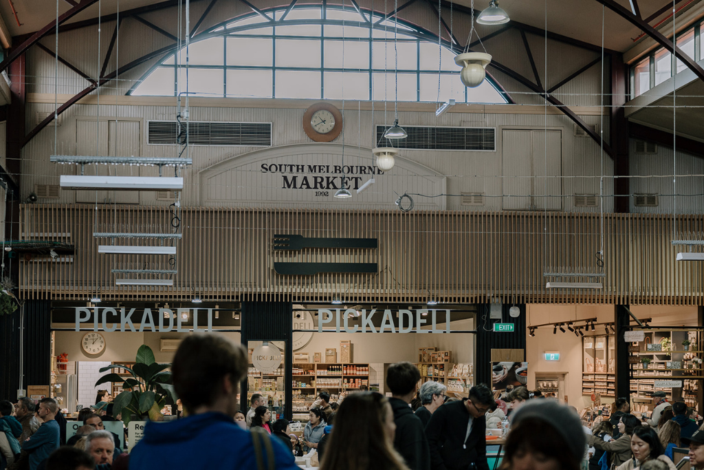Crowded indoor market with a sign reading 'South Melbourne Market' and a food stall named 'Pickadeli' amid shoppers and vendors.