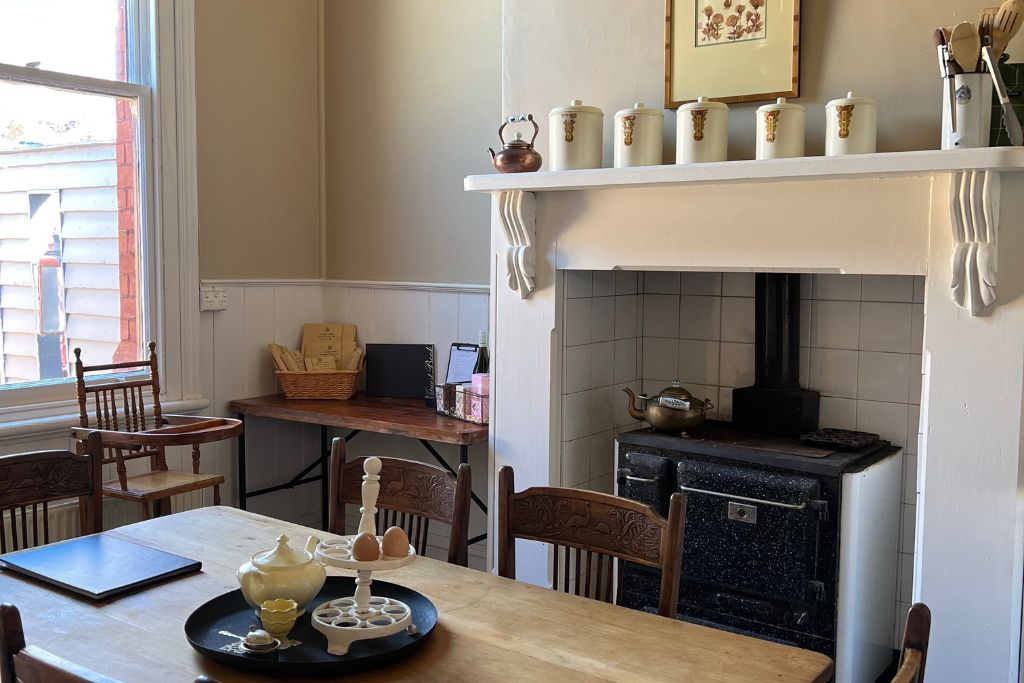 Interior of a cozy kitchen with a wood-burning stove, a wooden dining table with eggs and a teapot, and a window with natural light.
