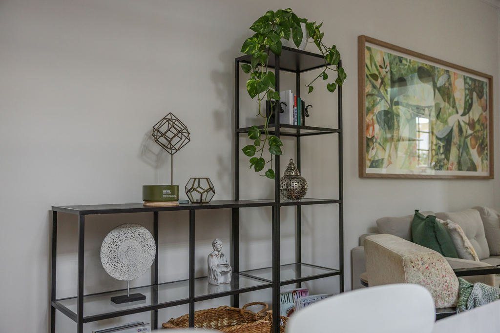 Decorative black metal shelving unit with green plant, books, and ornaments next to a beige couch and a colorful framed artwork on the wall.